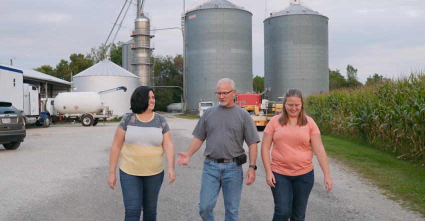 A man and two young women walk on a gravel road in front of a corn handling facility