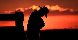 A farmer with his head down is silhouetted against a blazing orange sunset