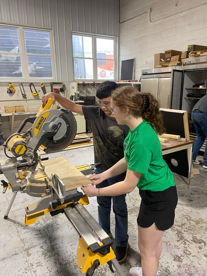 Hayley Gruenewald using a saw to cut plywood alongside a student