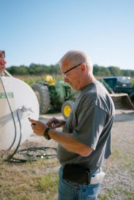 A man standing next to tractors and a big white tank while looking at his mobile phone