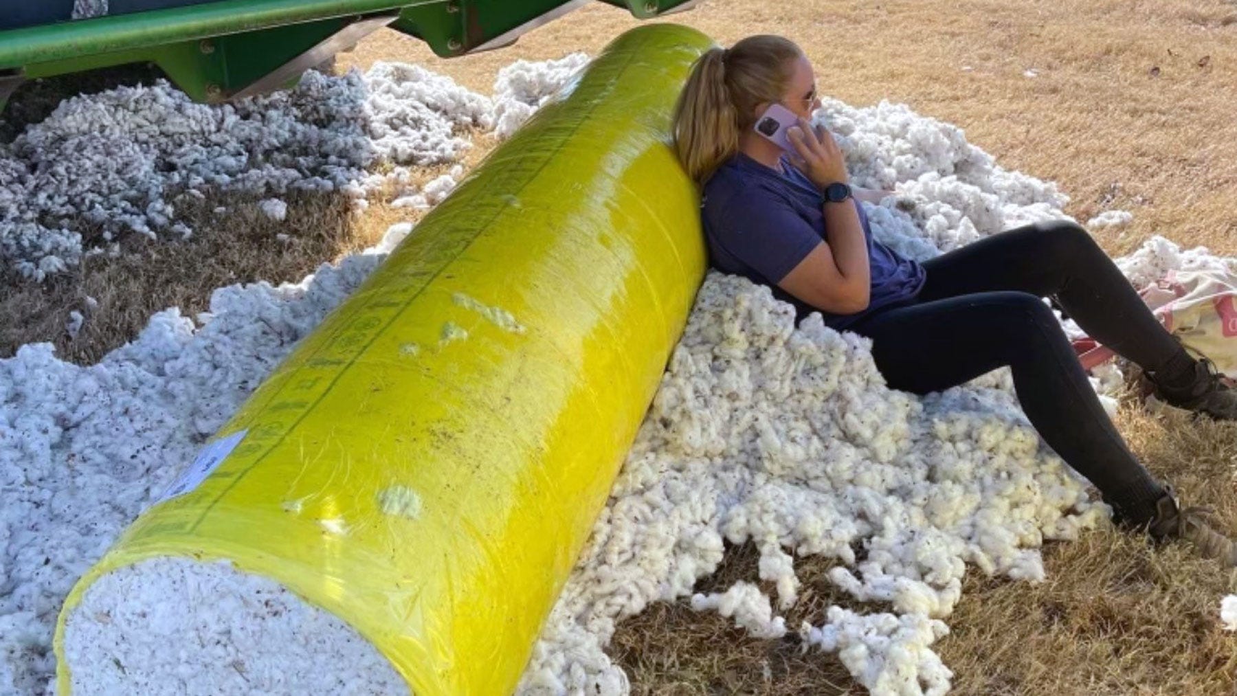 Woman sitting in pile of picked cotton beneath a parked cotton picker, leaned against a small cotton bale while taking a phone call.