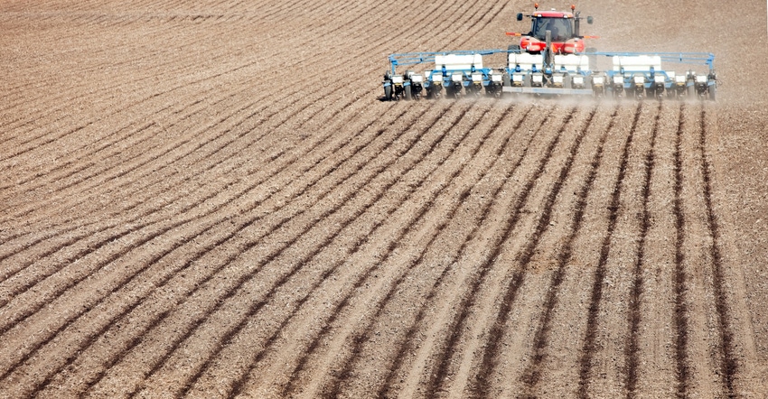 A low aerial view of a tractor planting a Spring cornfield. Focus is on the foreground soil, the tractor and planter are slig