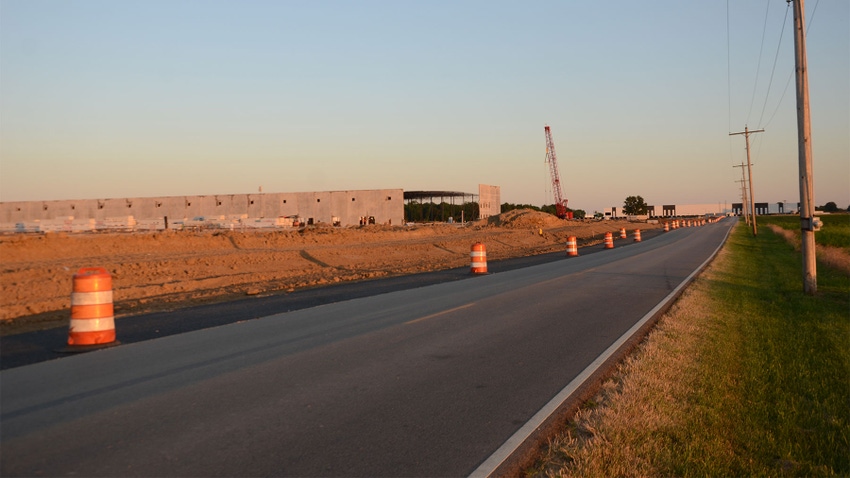 A paved road with farmland on the right side and a warehouse under development on the left side