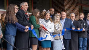 A group of men and young women in tiaras cut a blue ribbon in front of a building
