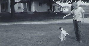 a young Tom Bechman plays with a dog in front of a farmhouse