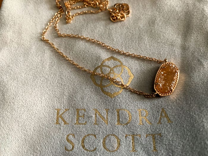 A gold neckless with a sparkly gem on a piece of cloth with a the Kendra Scott logo in gold embroidery