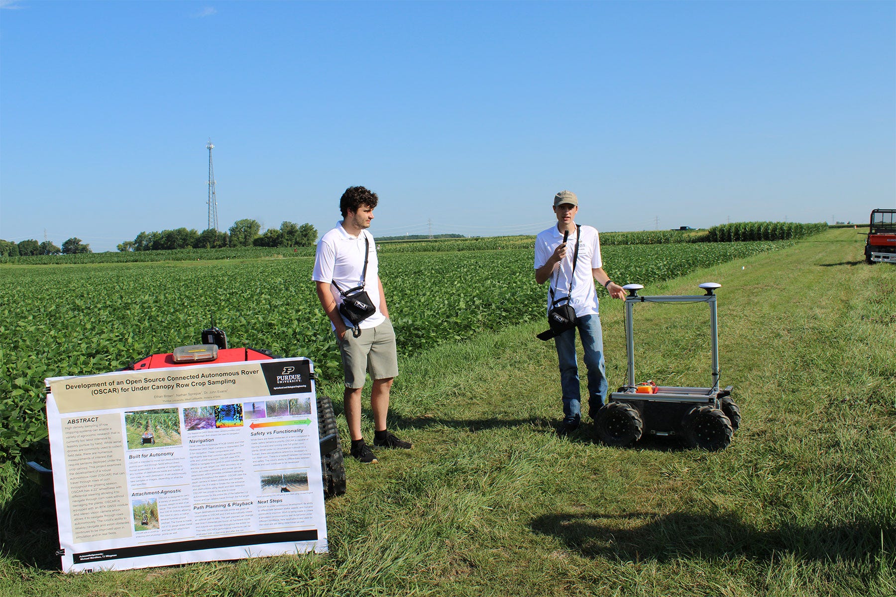 2 college students give a presentation with a robot in front of a soybean field