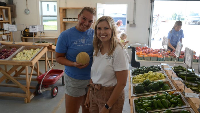 Two women posing for a photo at a market while one holds a cantaloupe Two women posing for a photo at a market while one holds a cantaloupe