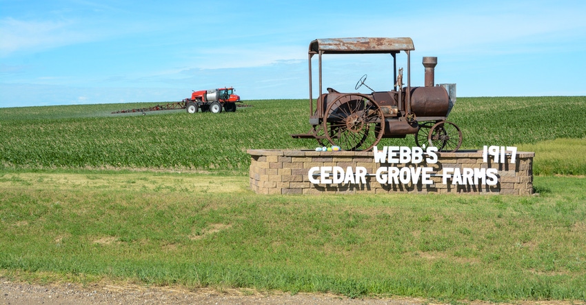 The Cedar Grove Farm sign and a tractor spraying a field in the background