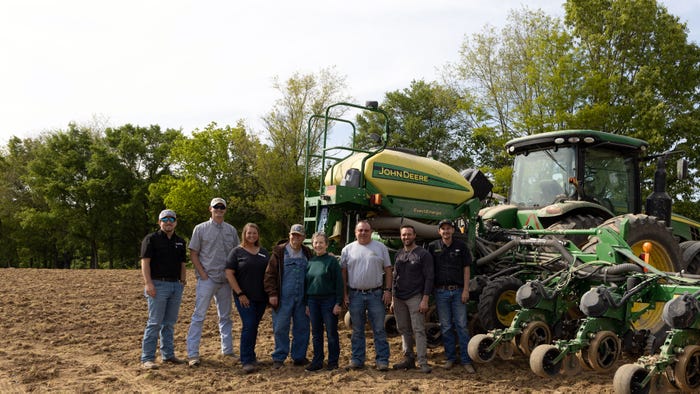 Group of 8 people standing in front of a planter hooked to a tractor in a field