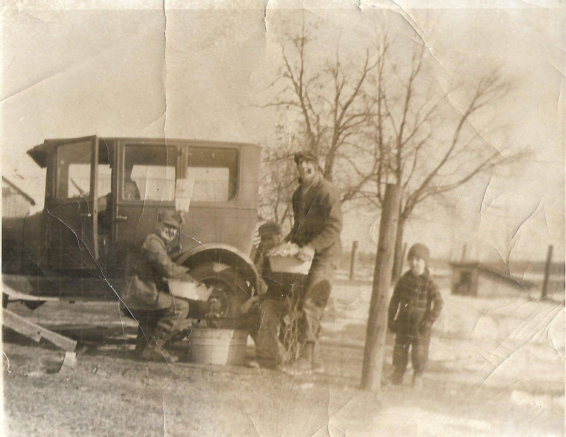A worn, sepia-toned photograph of three men and a boy working with a meat grinder attached to a Model T Ford