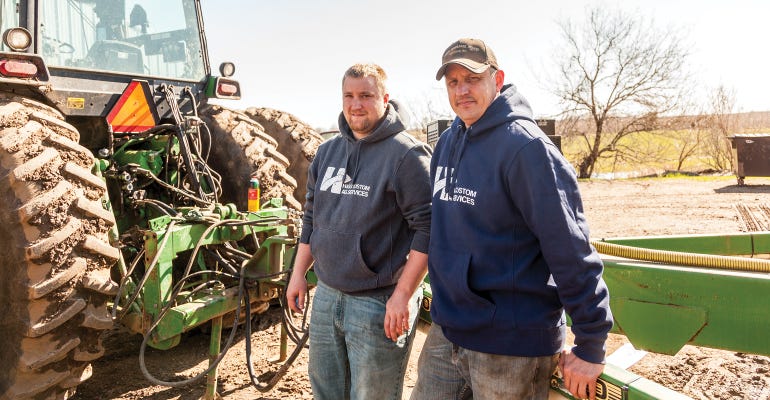Two men standing between a John Deere rear tractor tire and implement.