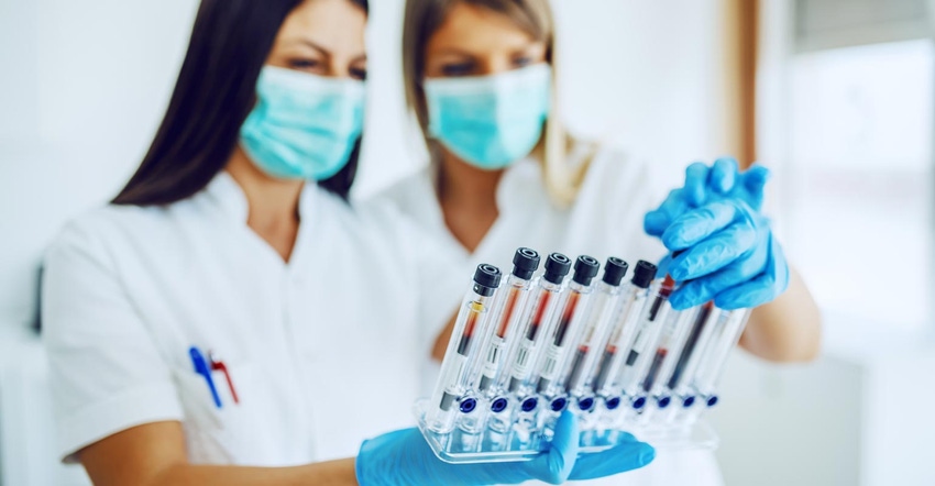 Two women in white lab coats wearing blue medical gloves, hold a tray of test tubes