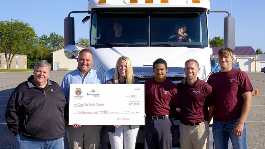 A group of adults and students smile as they pose in front of a semitruck holding a large check 