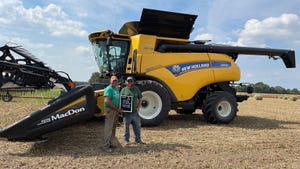 Terry Hayhurst and son-in-law Kegan Knust hold a sign and smile in front of a yellow combine in a harvested field
