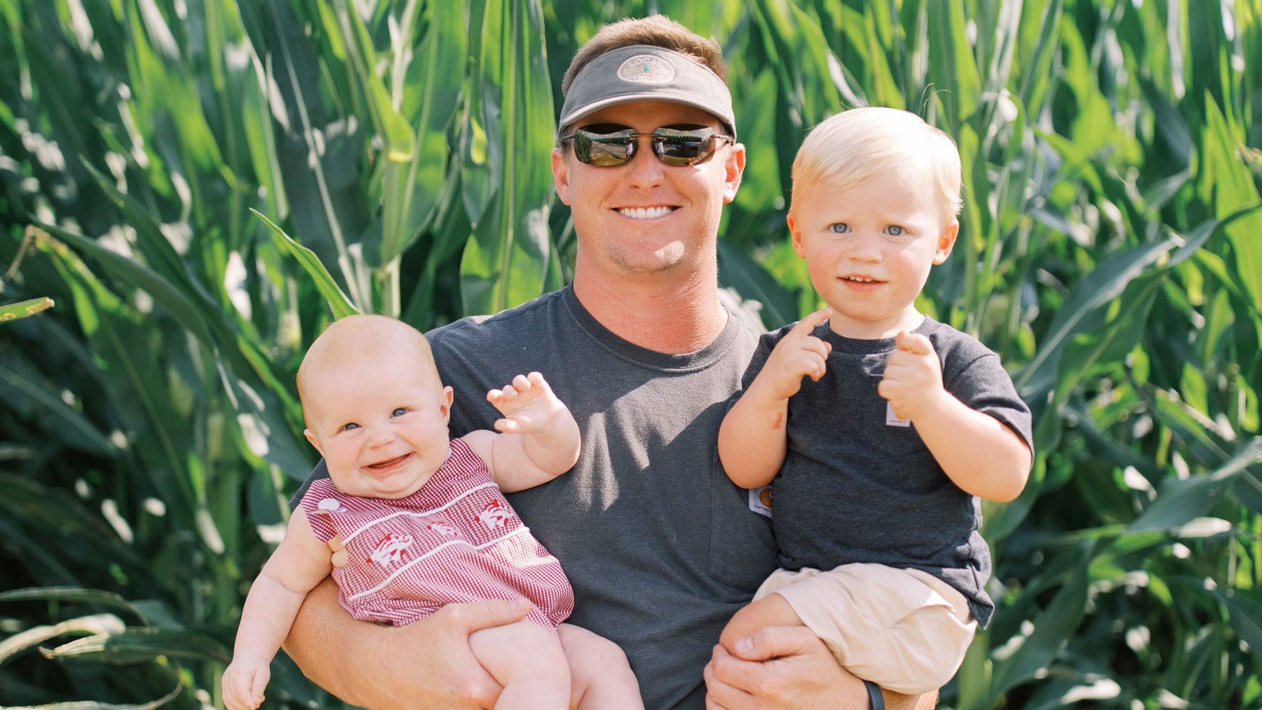 Father holding infant son and toddler son, standing in front of a corn field.