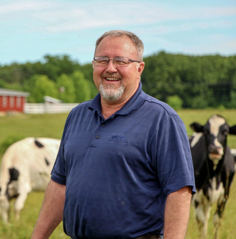 Farmer Jack Dill smiles as he poses for a photograph with a red barn and dairy cows in the background