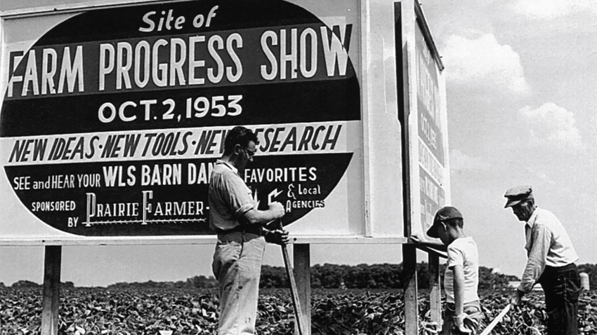 A black-and-white photograph of workers installing a sign for the Farm Progress Show