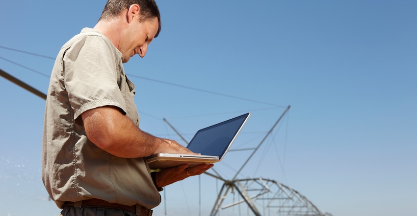 Farmer standing in front of crop irrigation equipment working on laptop