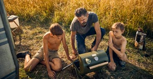 Father with two boys operating a small grill while out camping