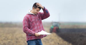 Worried young farmer standing on field and  looking at papers from bank, tractor in background