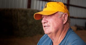 A close up of a farmer sitting in a barn wearing a yellow cap and looking off into the distance
