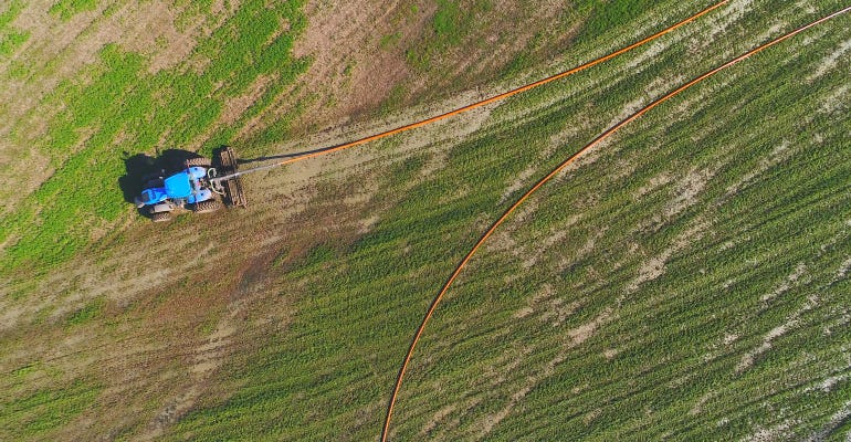 Aerial view of tractor fertilizing field with liquid manure pumped from remote tanker truck via giant hose