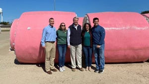 Five people standing in front of round module cotton bales wrapped in pink plastic.