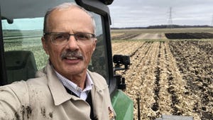 A man with glasses takes a photo of himself on a tractor with a field in the background