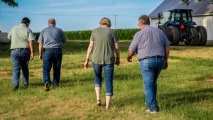 a woman and three men walking away, toward a cornfield and blue tractor