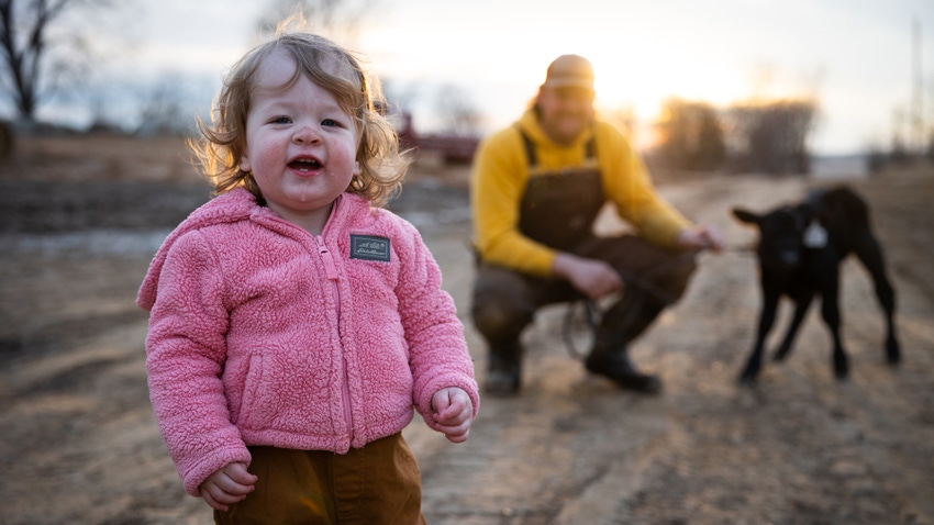 Toddler Clare Haynes in foreground wearing pink sherpa coat with Dan Haynes kneeling with black baby calf out of focus behind Toddler Clare Haynes in foreground wearing pink sherpa coat with Dan Haynes kneeling with black baby calf out of focus behind
