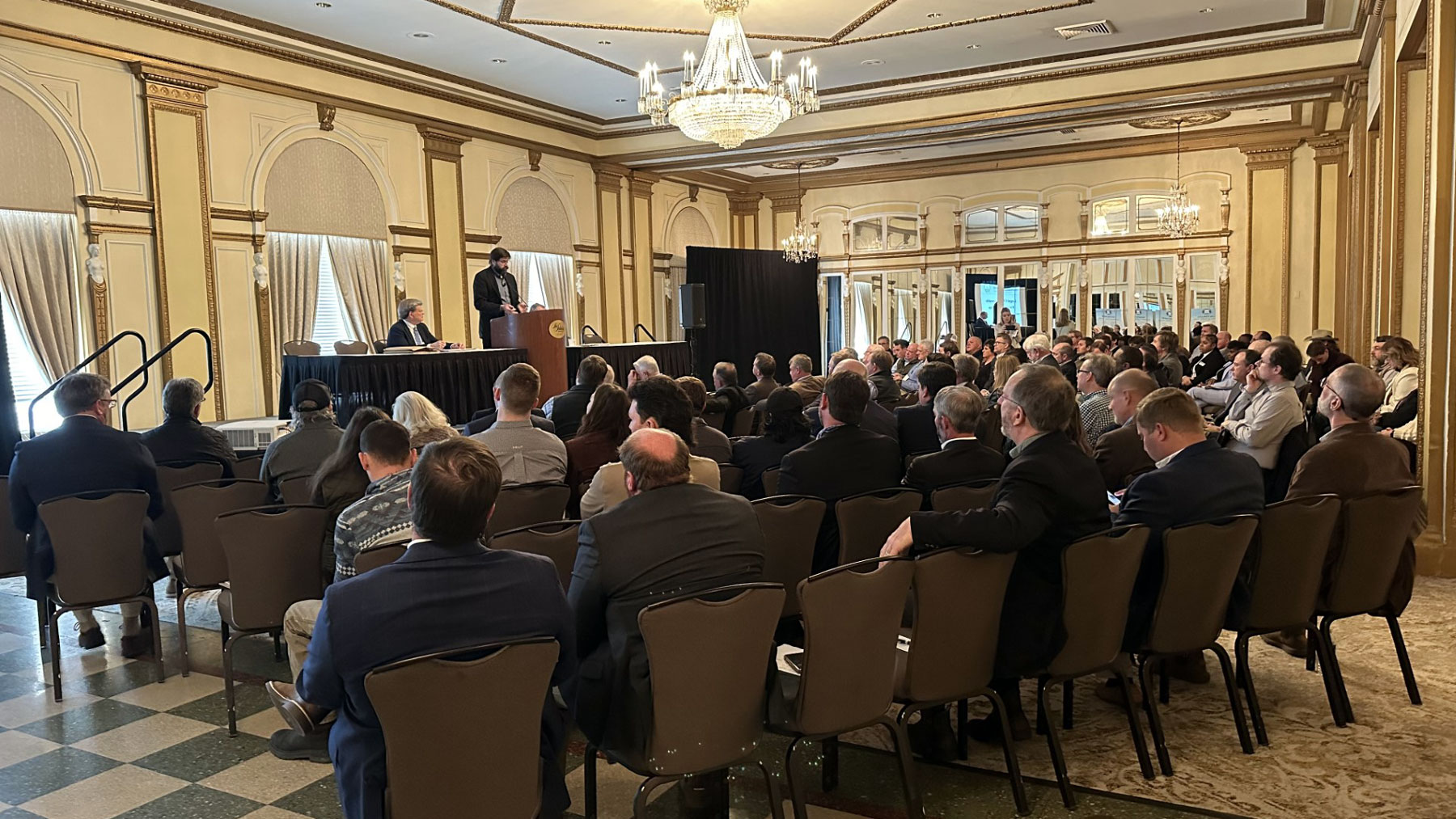 Man presenting on stage to a large group of people at a formal meeting.
