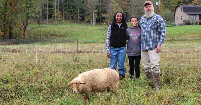 A husband and wife stand with their daughter as they smile at the camera and a sheep grazes past them