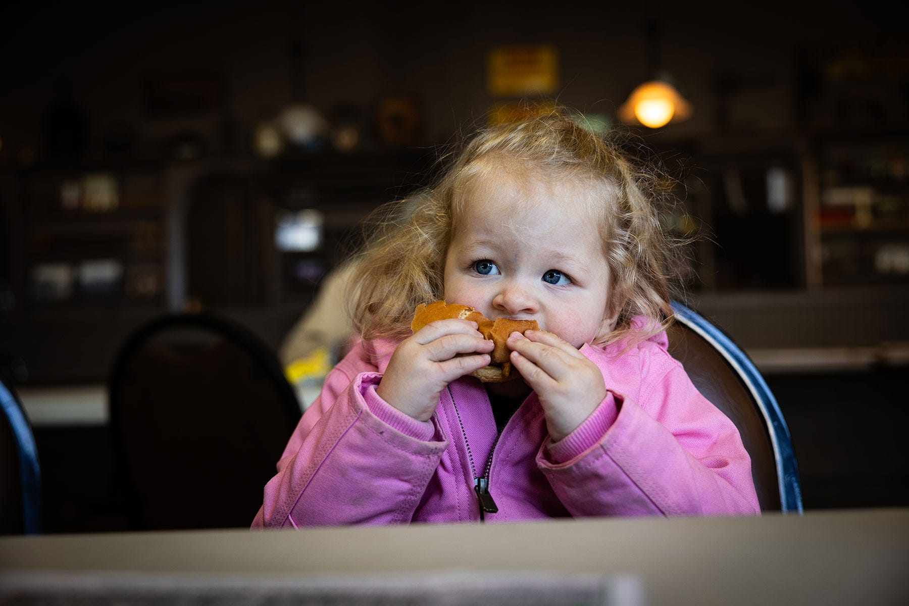 A young girl wearing a pink coat and eating a cheeseburger