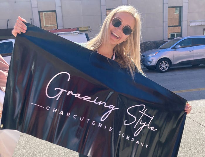 Rachel Holkenbrink smiles while holding a sign for her Grazing Style Charcuterie Company