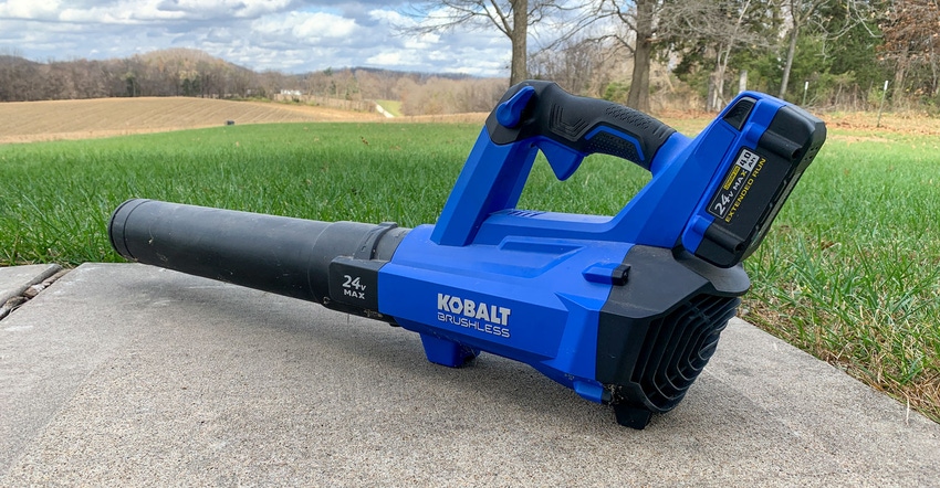 A cobalt blue and black leaf blower placed on a patch of concrete next to pasture