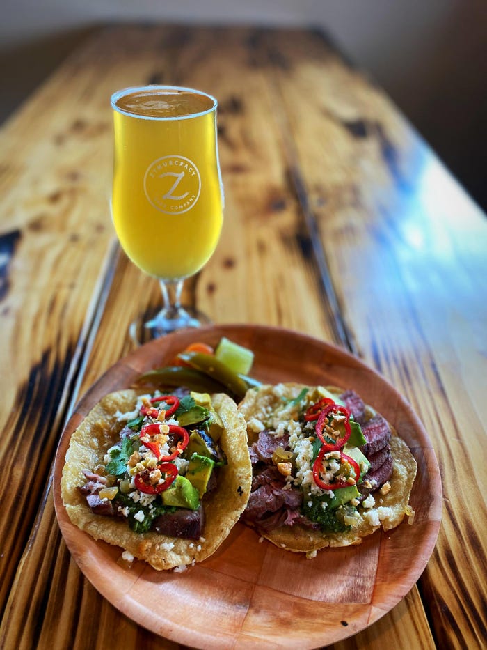 Two steak tacos with colorful toppings on a bamboo plate and a glass of a light yellow beer