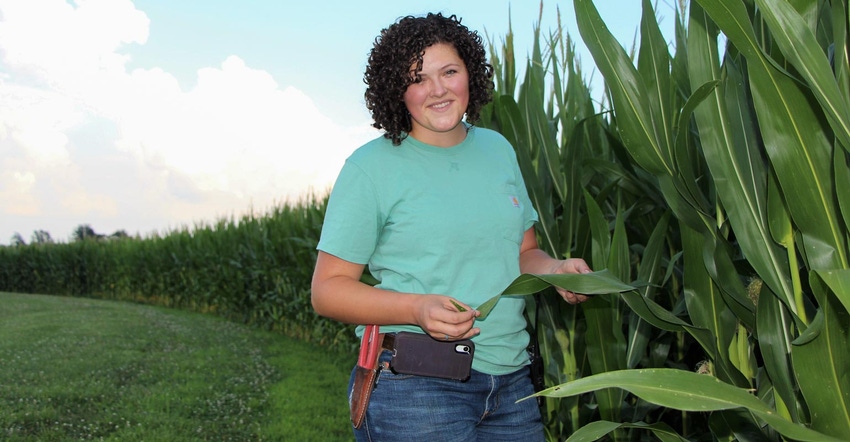 A young lady standing next to a corn field, holding a corn leaf and smiling as she poses for a photo