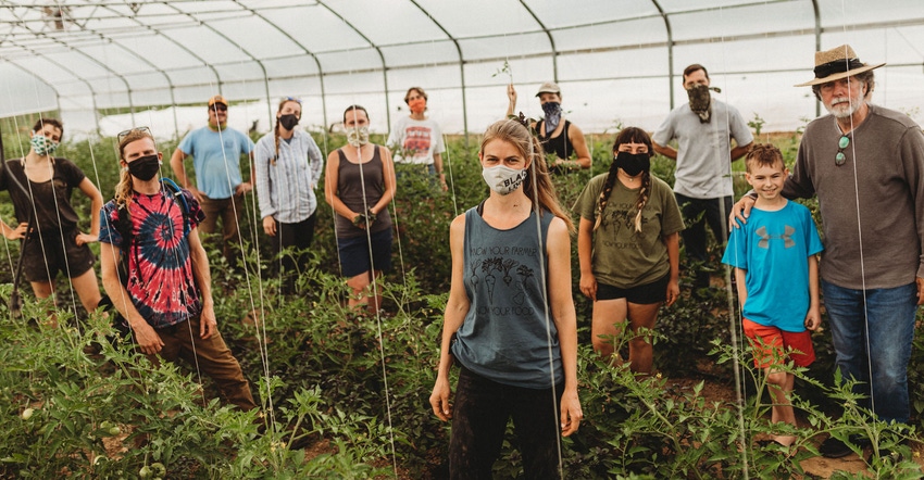 A group of various people, wearing face masks, stand between rows of crops in a greenhouse