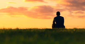 silhouetted solitary man seated in grass and looking into sunset