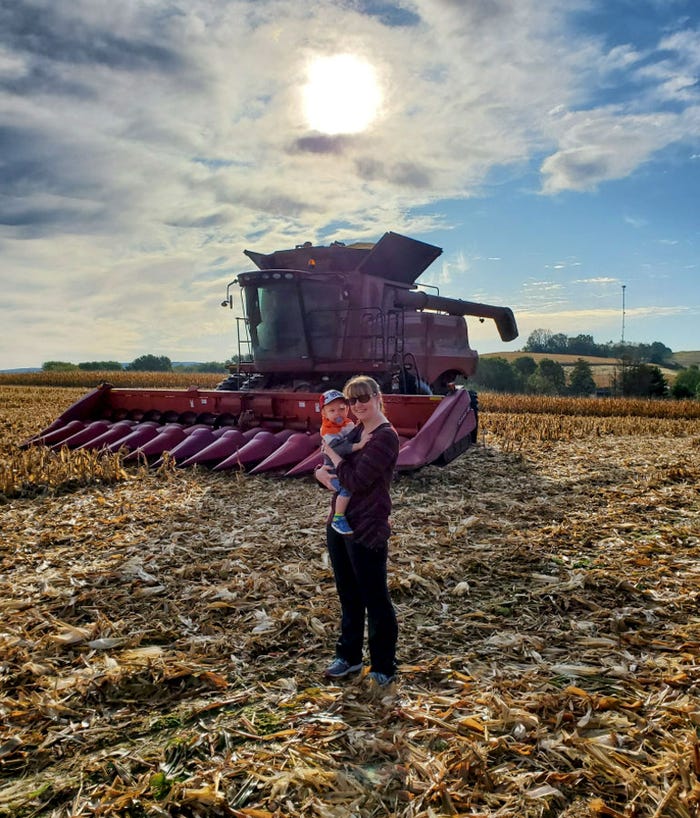 Sheilah Reskovac stands in a partly-harvested corn field holding her son with the combine behind them