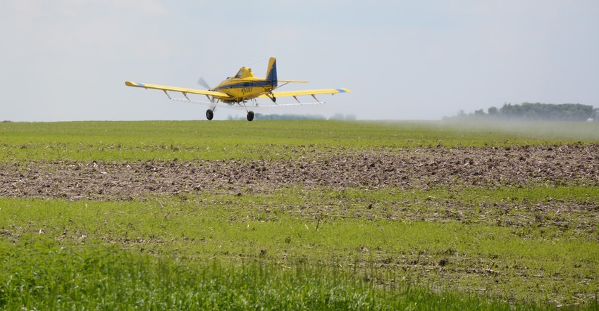 a field being aerial sprayed by a small yellow plane flying across blue sky