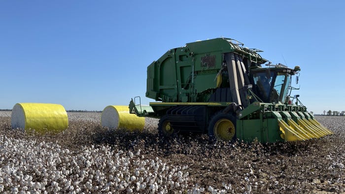 Cotton baler picker harvesting cotton field with two yellow wrapped cotton modules behind it.