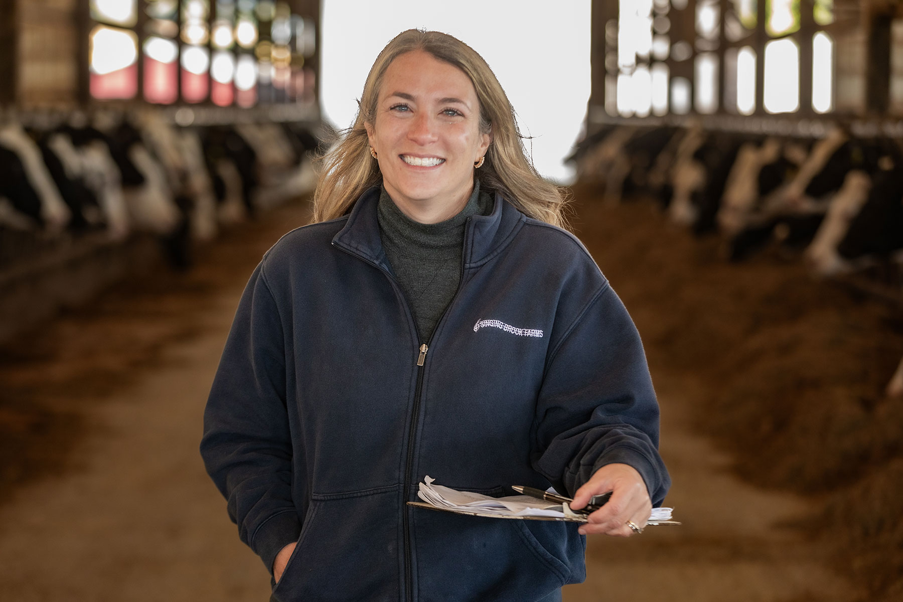Woman holding a clipbard smiles as she stands in a dairy barn