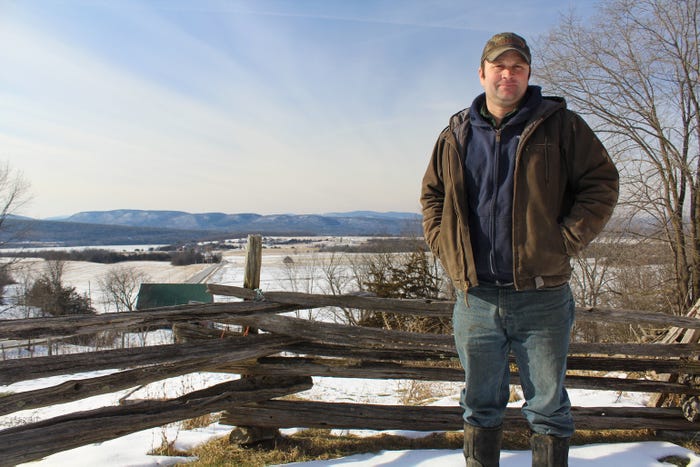 Farmer Jon Lucas stands near a fence with a scenic view of fields and mountains behind him in Orwell, Vt.