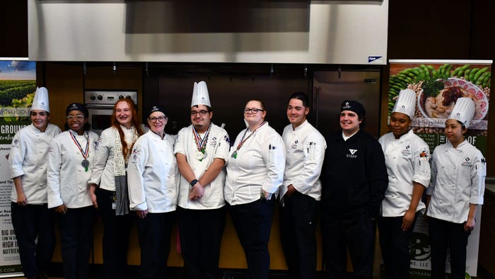 Group of people wearing chef suits and hats standing in a demonstration kitchen.