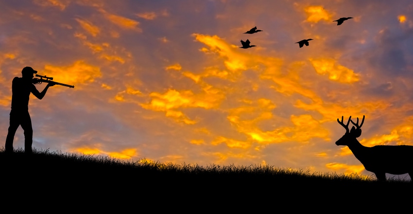 Silhouette of a hunter aiming at a buck against an evening sunset.