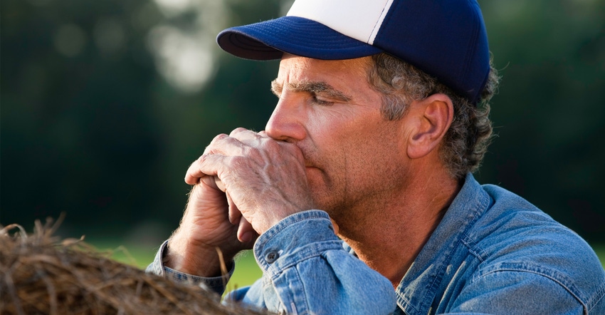 "Worried adult farmer leaning on hay bale, praying with eyes closed"