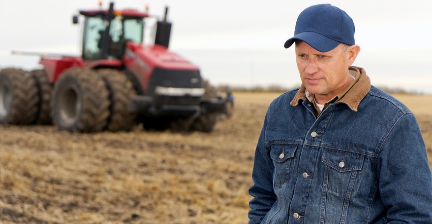 A discouraged farmer in a field in front of a tractor.