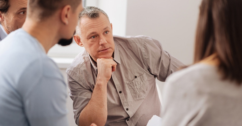 Smart positive middle-aged male holding elbow on the right leg sitting in the office while having conversation with his cowor